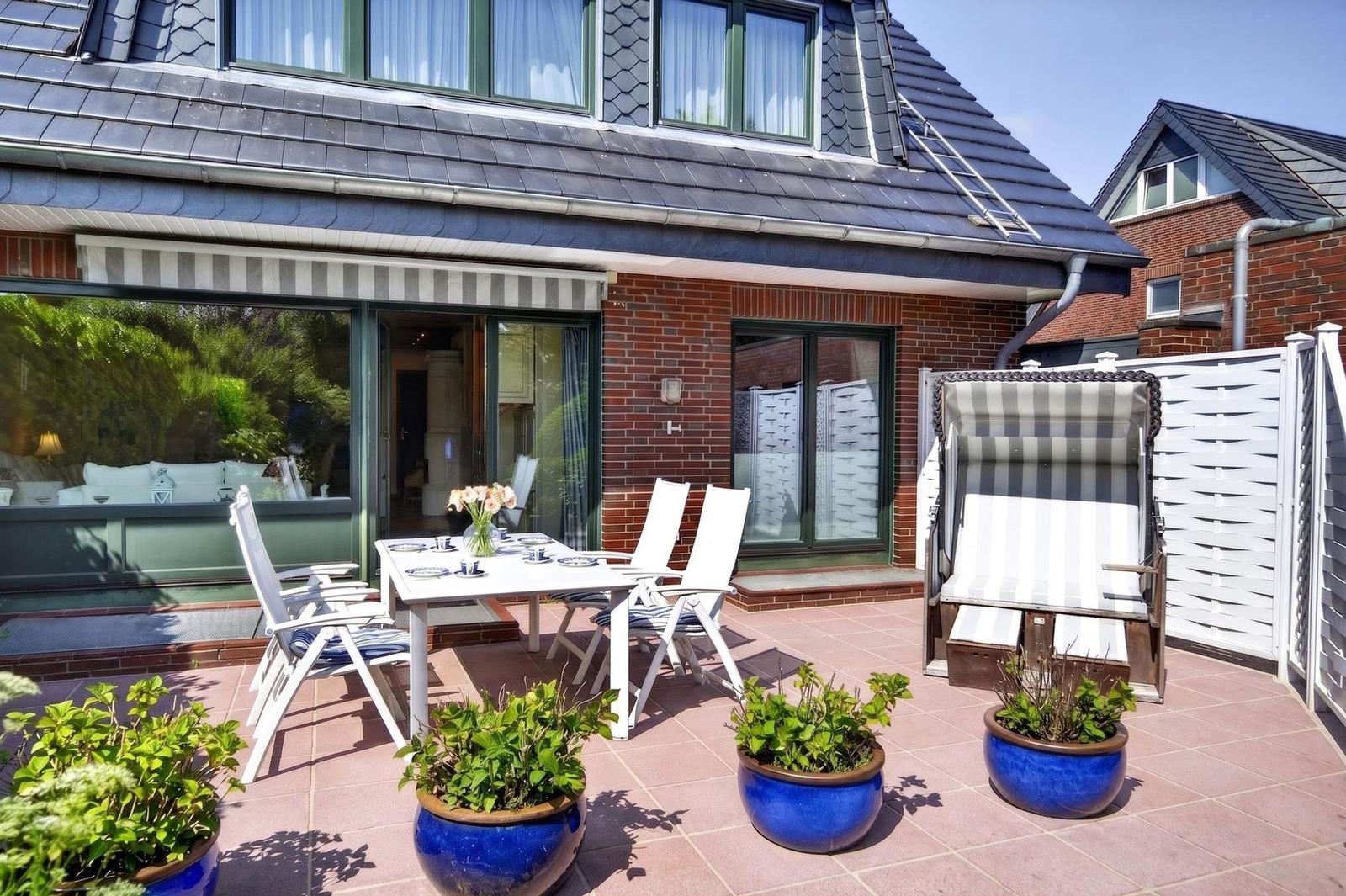 Terrasse mit Tisch, Stühlen und blauen Pflanzkübeln. Hintergrund: Haus mit Glasfront und Dach.