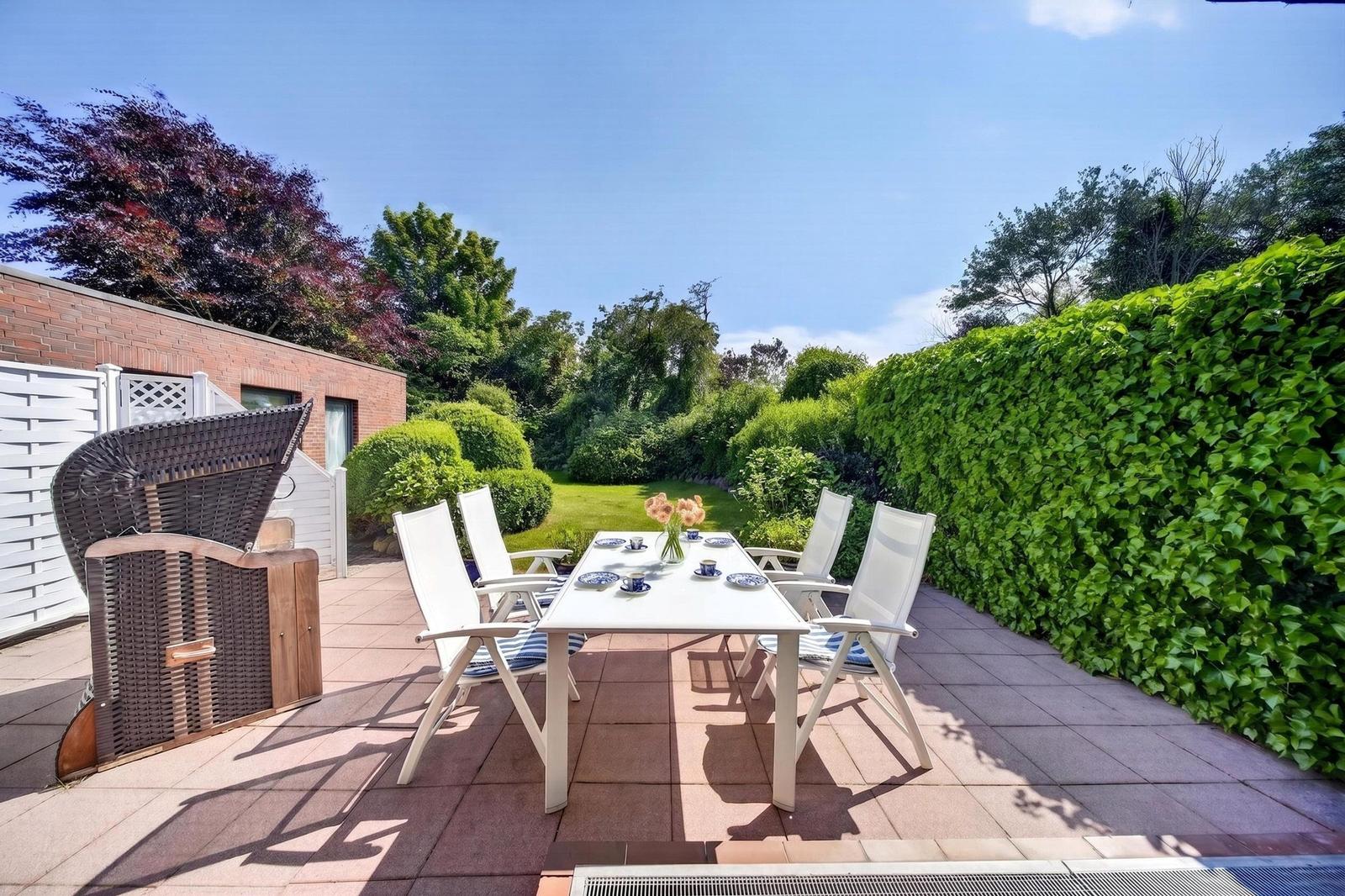 Terrasse mit weißem Tisch und Stühlen, umgeben von grünen Hecken und einem Garten.