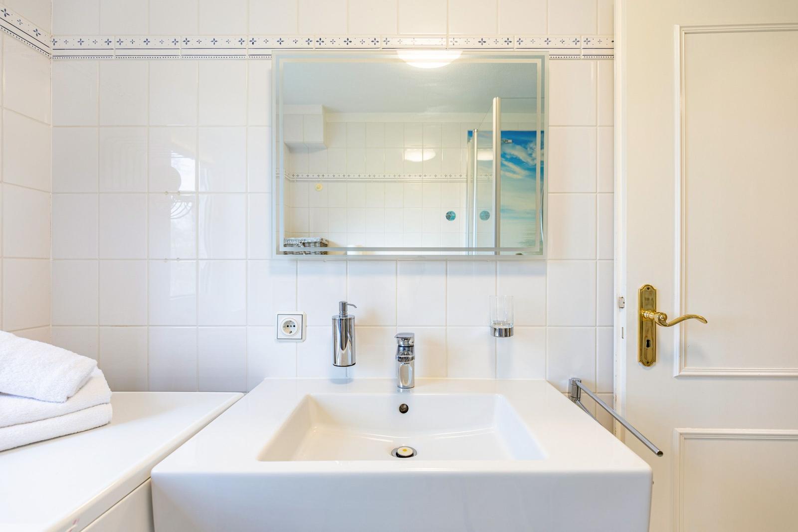 Bathroom with sink, mirror, and shower. White tiles and gold handle.