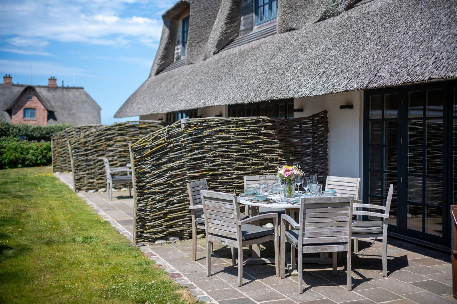 Terrace with table and chairs in front of a house with thatched roof and willow fence.