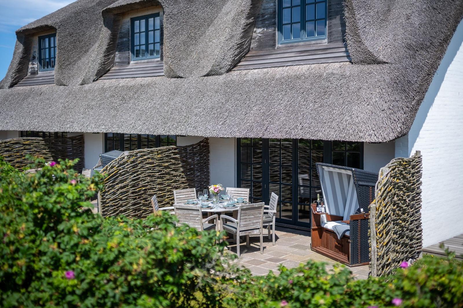 Terrace with table, chairs, and lounge under thatched roof. Surrounded by plants and wooden fence.