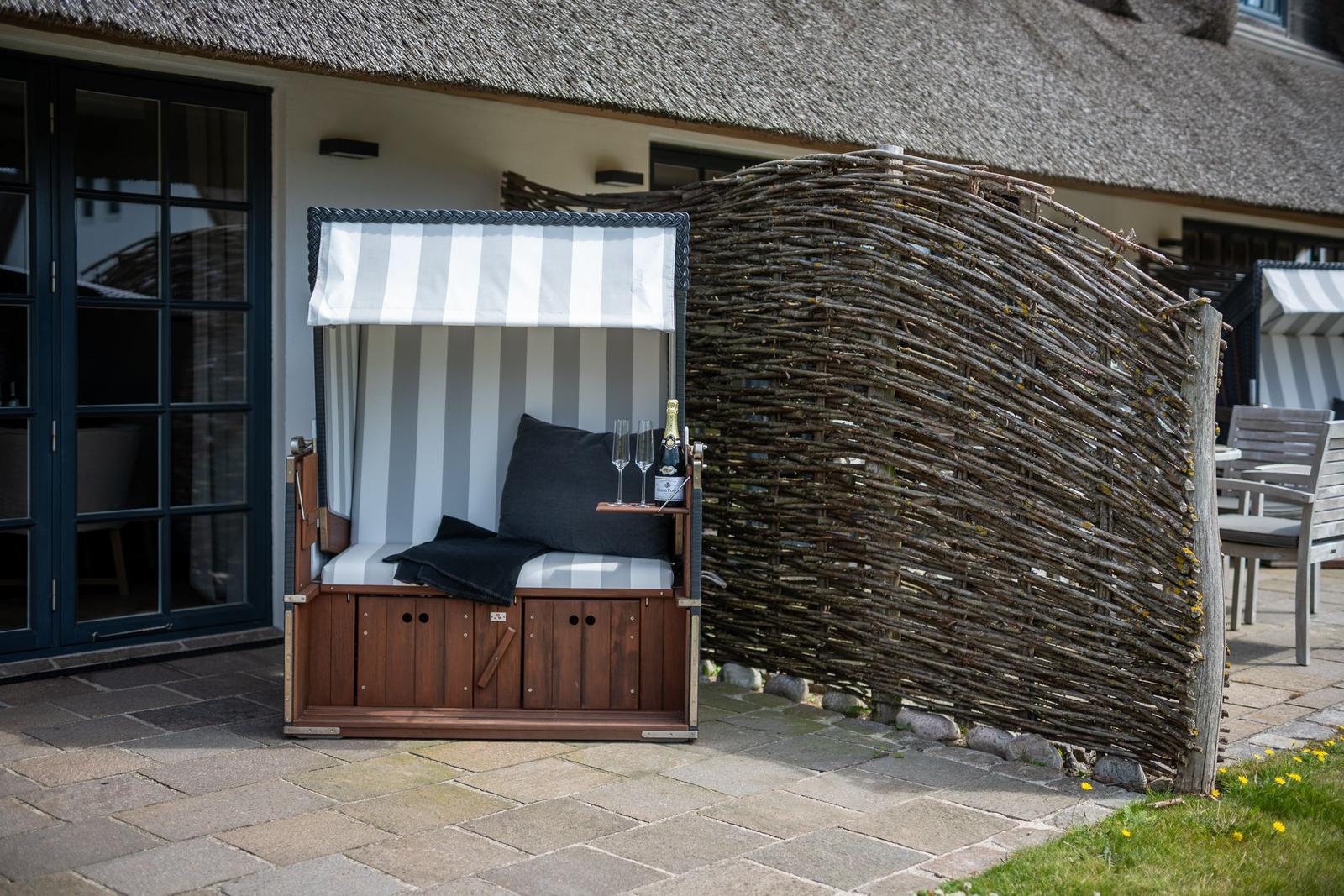 Hütte mit Streifenmarkise, Sitzfläche, Kissen und Flasche Champagner. Hintergrund: Holzschirm und Terrasse.