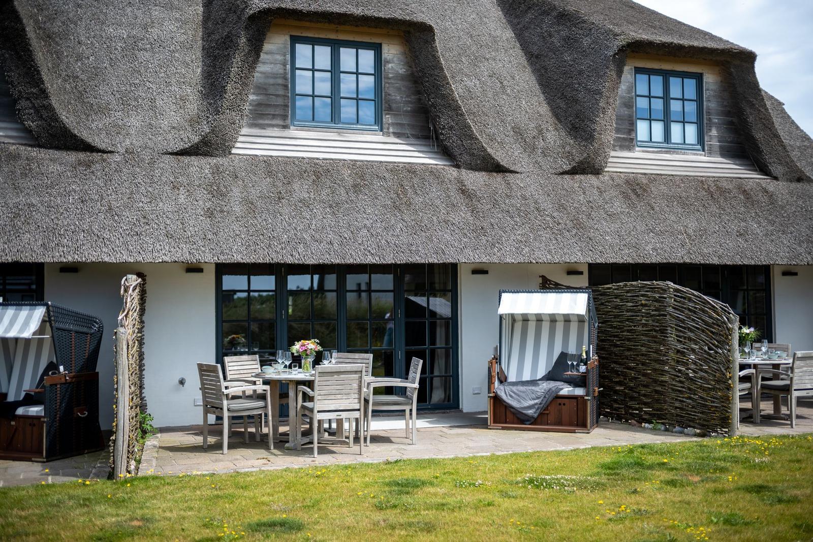 Haus mit Strohdach, Terrasse mit Tisch, Stühlen und Strandkörben.