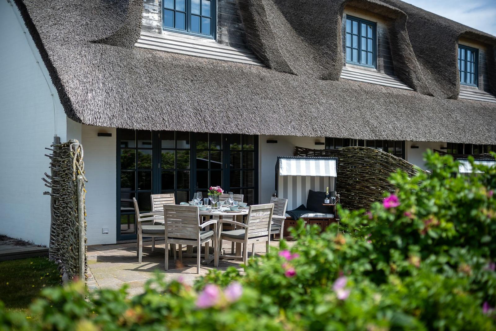 Terrasse mit Tisch und Stühlen vor einem Haus mit Strohdach und Fenstern.