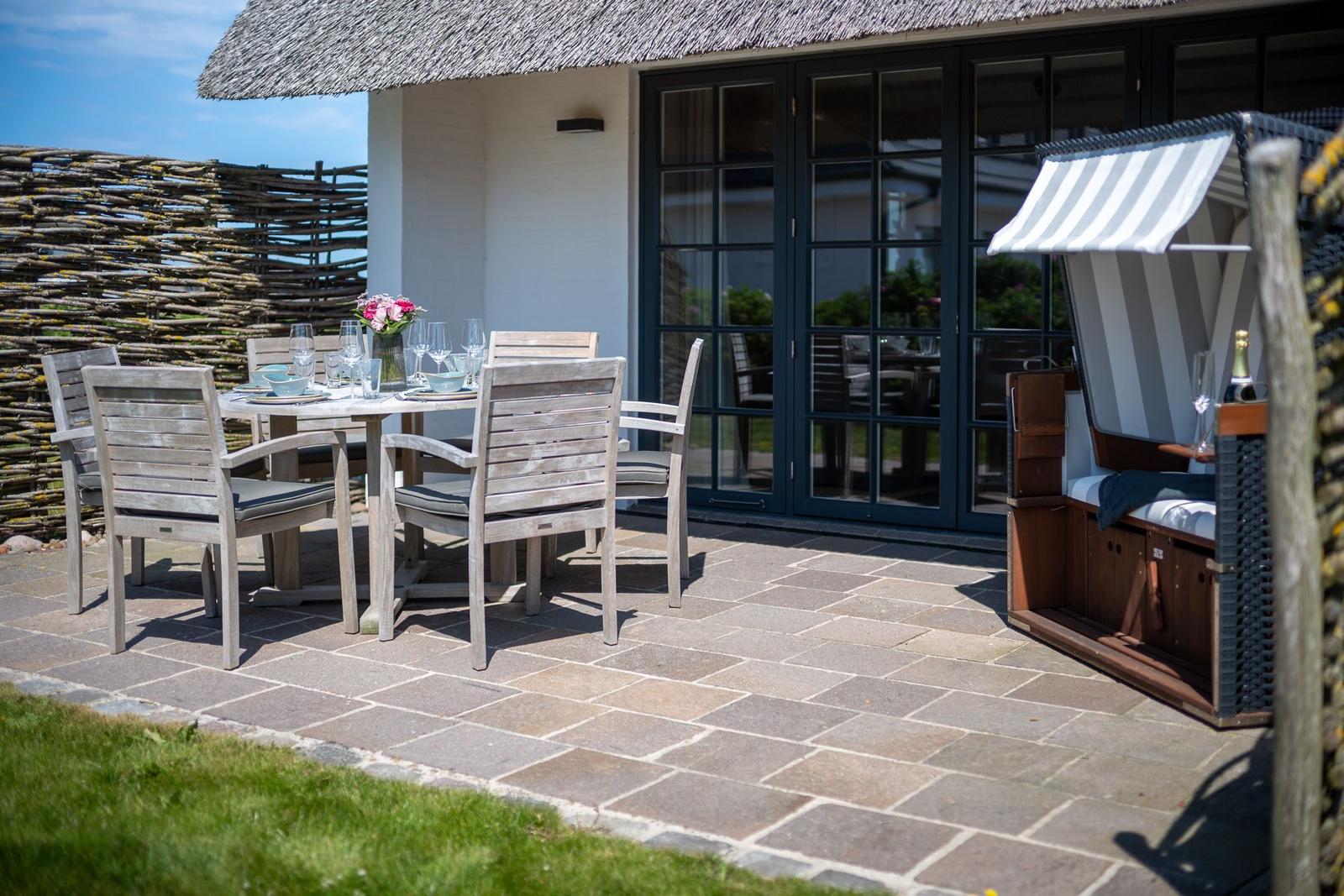 Terrasse mit Tisch, Stühlen und Strandkorb. Hintergrund: Haus mit Schiebetüren und Strohdach.