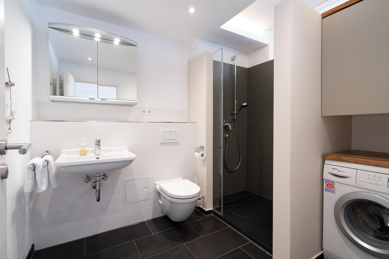 Badezimmer mit Dusche, Waschbecken und Toilette. Nebenraum mit Waschmaschine.