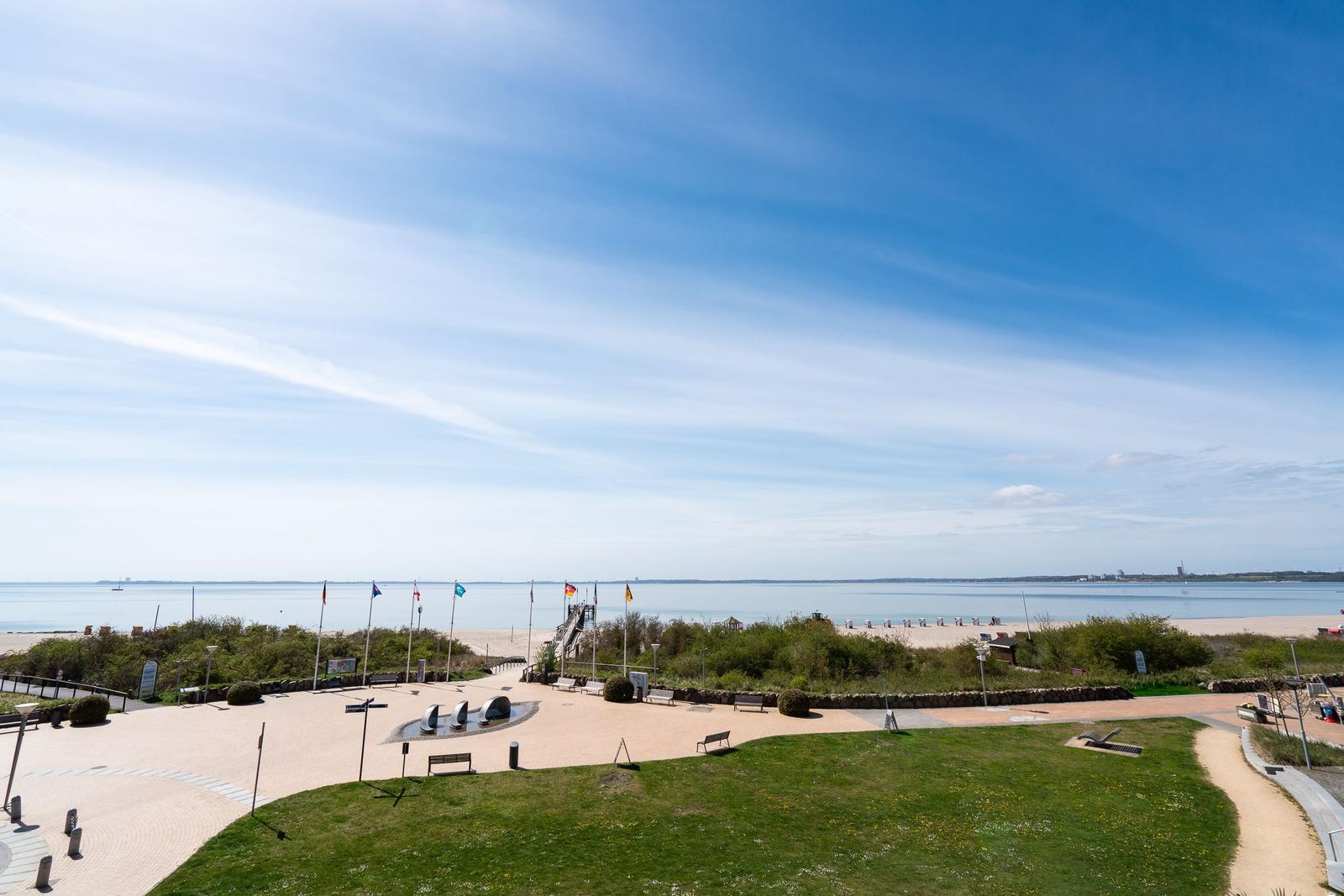Weitläufiger Strandbereich mit Grünflächen, Bänken und Flaggen unter blauem Himmel.