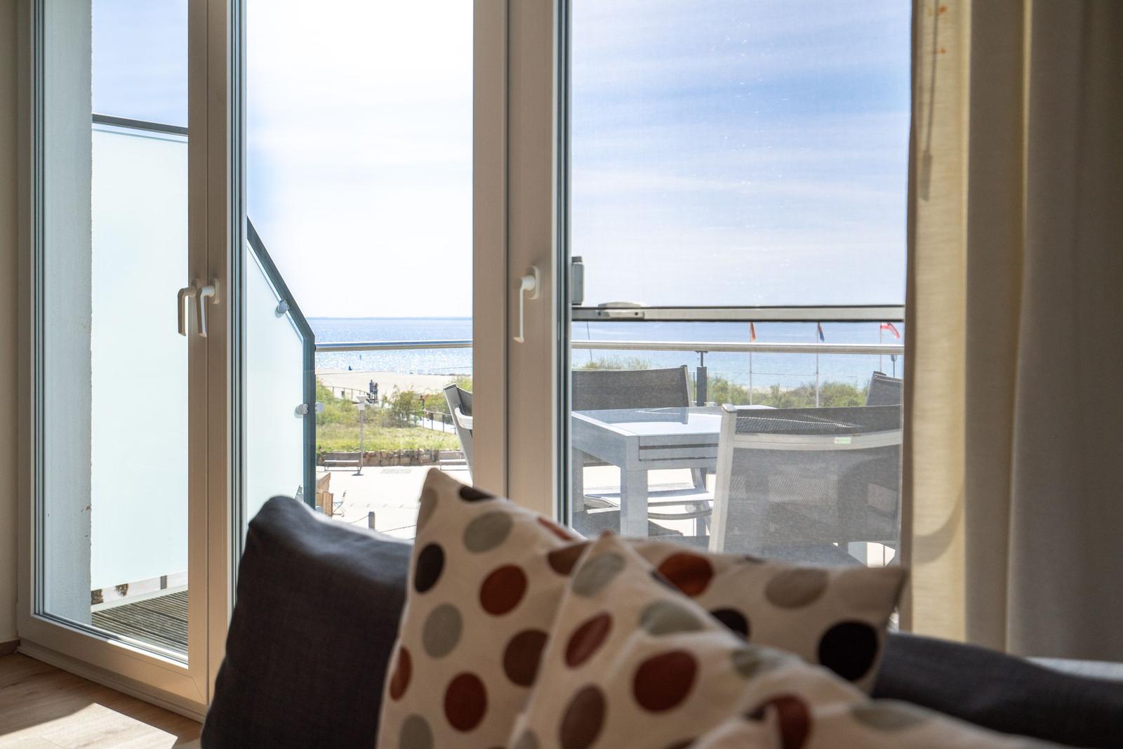 Durch das Fenster ist eine Terrasse mit Tisch und Stühlen sowie Blick aufs Meer zu sehen.