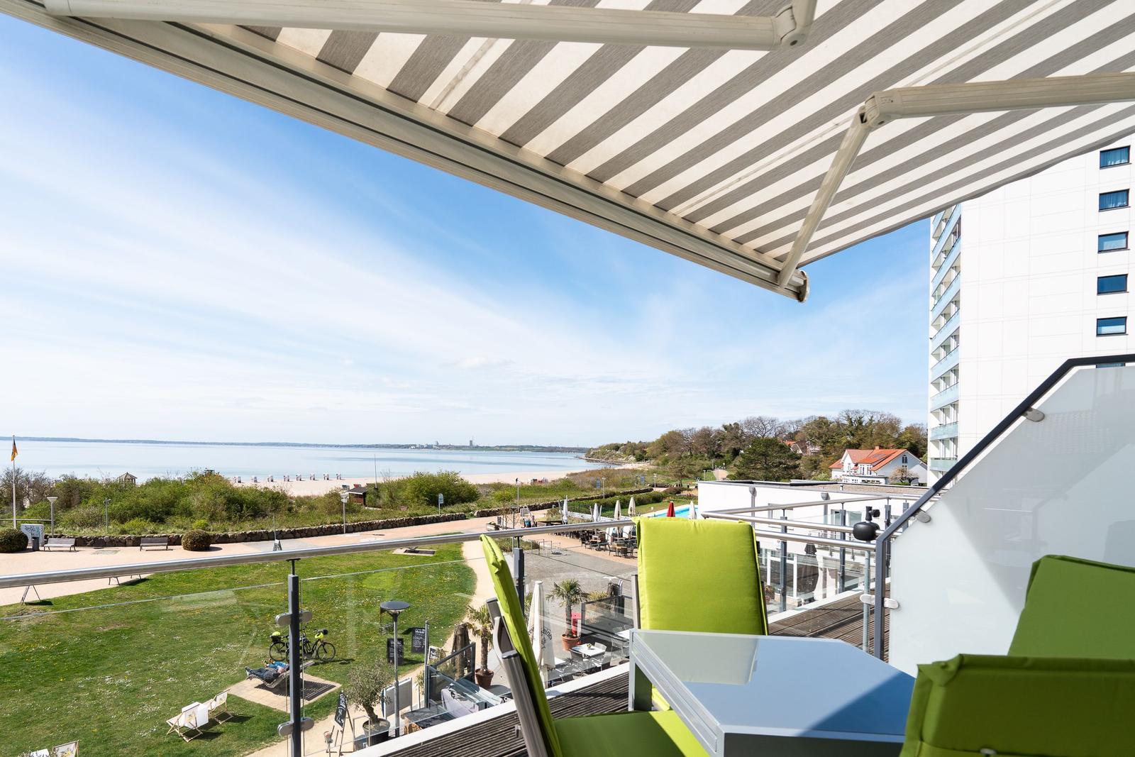 Terrasse mit Sonnenschutz, grünen Stühlen und Blick auf Strand und Wasser.