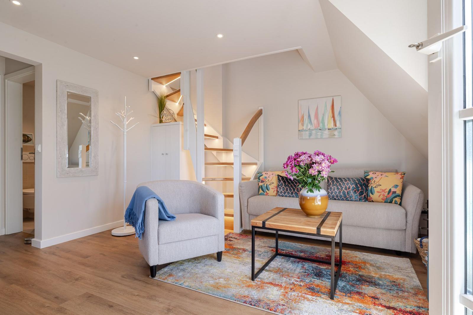 Living room with sofa, armchair, table, and flowers. Staircase and mirror are visible.