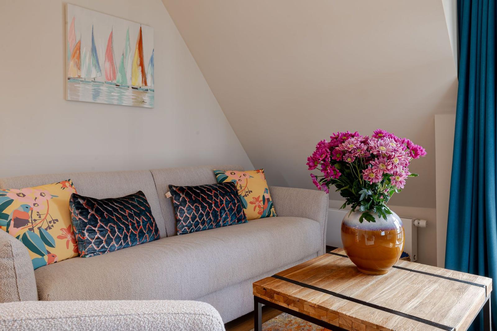 A sofa with cushions is under a sloped ceiling. A table with flowers and a painting are beside it.