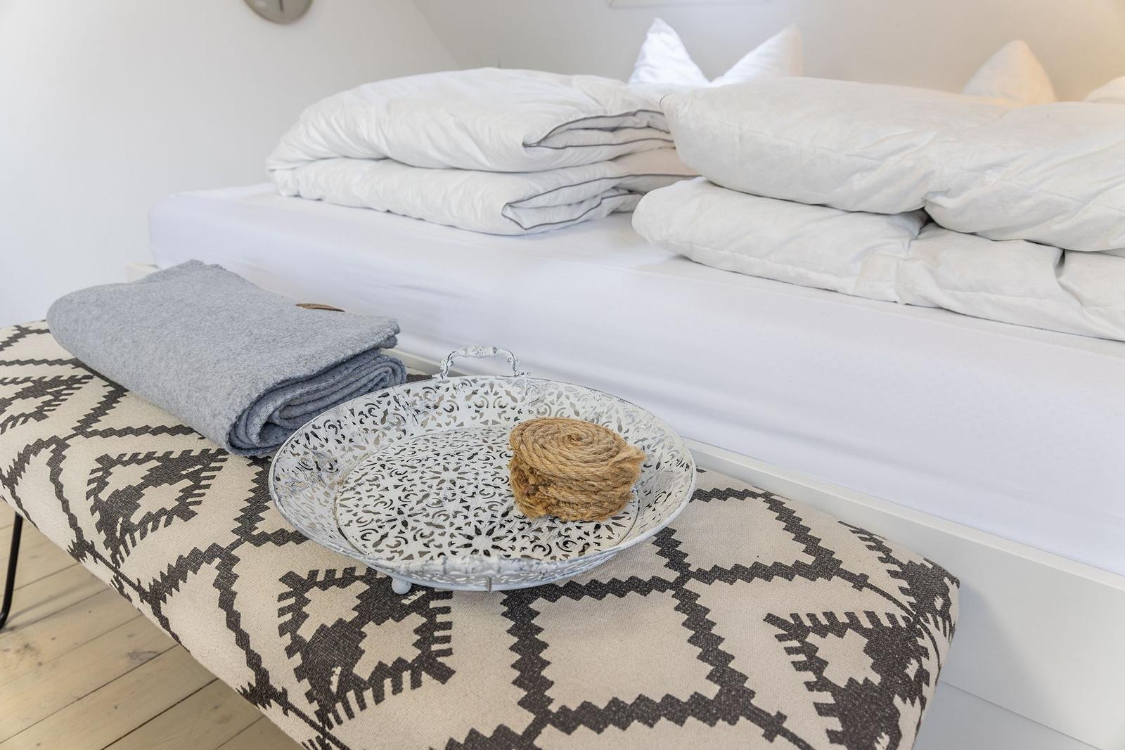 Ein Bett mit weißen Laken und Kissen. Davor steht ein Hocker mit Muster und einer Schale.
