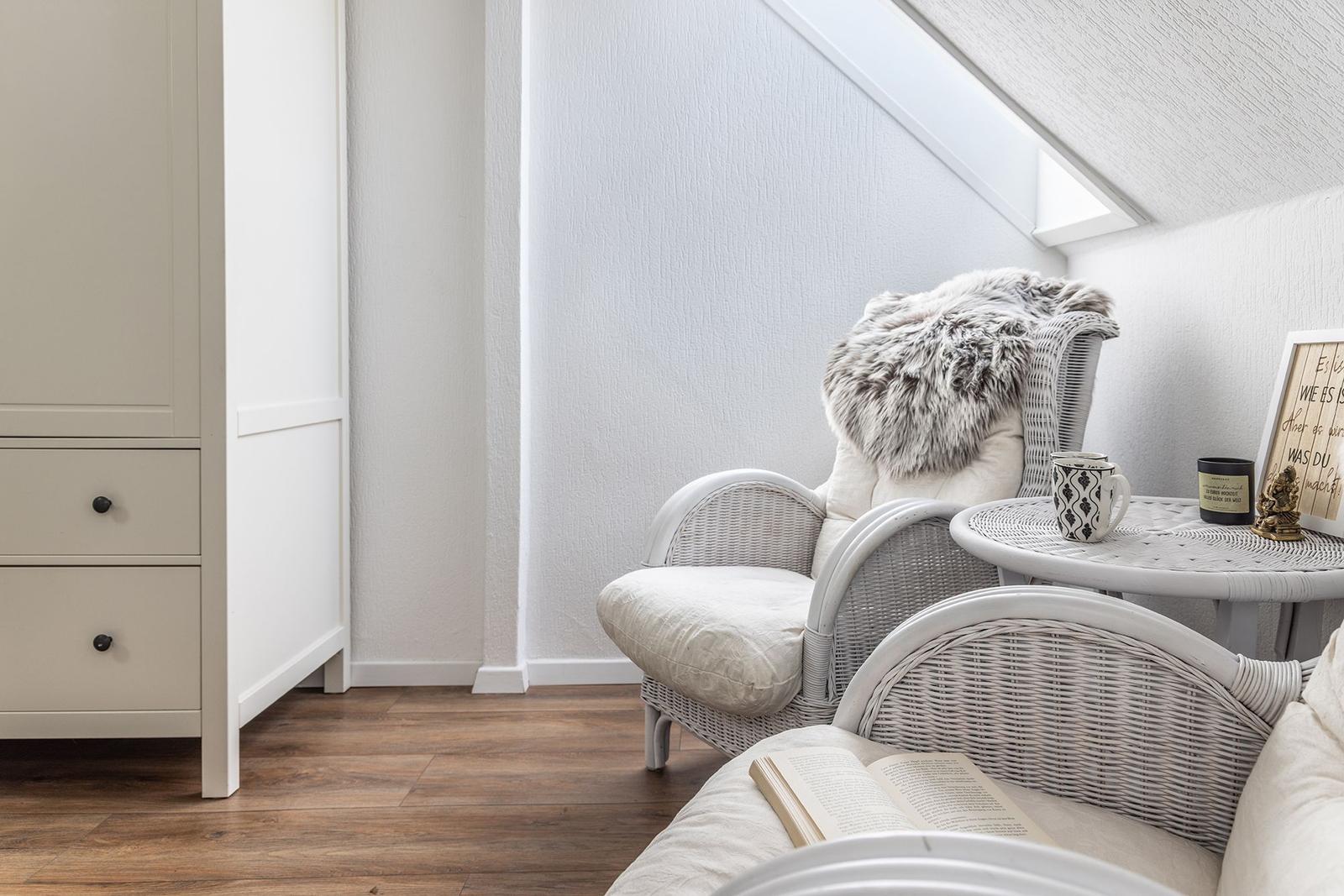 Wohnbereich mit weißen Rattan-Stühlen, kleinem Tisch und Holzboden unter Dachschräge.