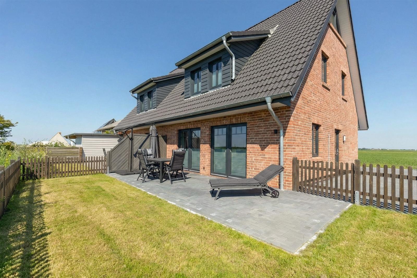 Modernes Haus mit Terrasse, Grillplatz und Liege. Umgeben von Grünflächen und Holzzaun.