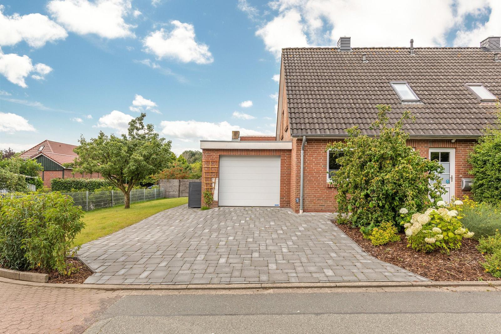 Haus mit Garage, gepflastertem Vorplatz und grüner Umgebung unter blauem Himmel.