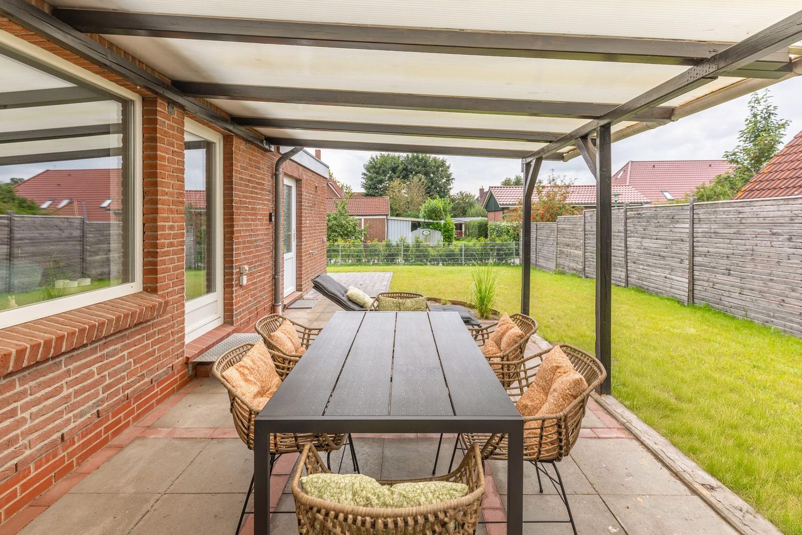 Terrasse mit Tisch und Stühlen unter Dach, Grasfläche und Zaun im Hintergrund.