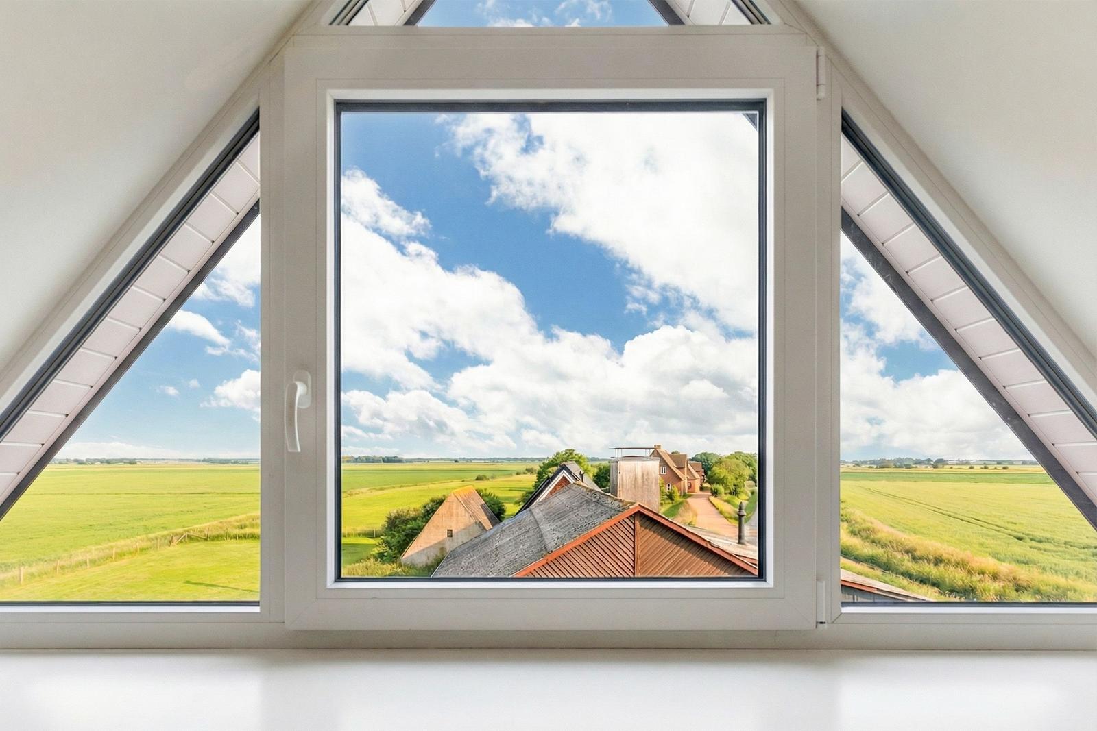 Dachfenster mit Blick auf grüne Felder und Dächer unter blauem Himmel mit weißen Wolken.