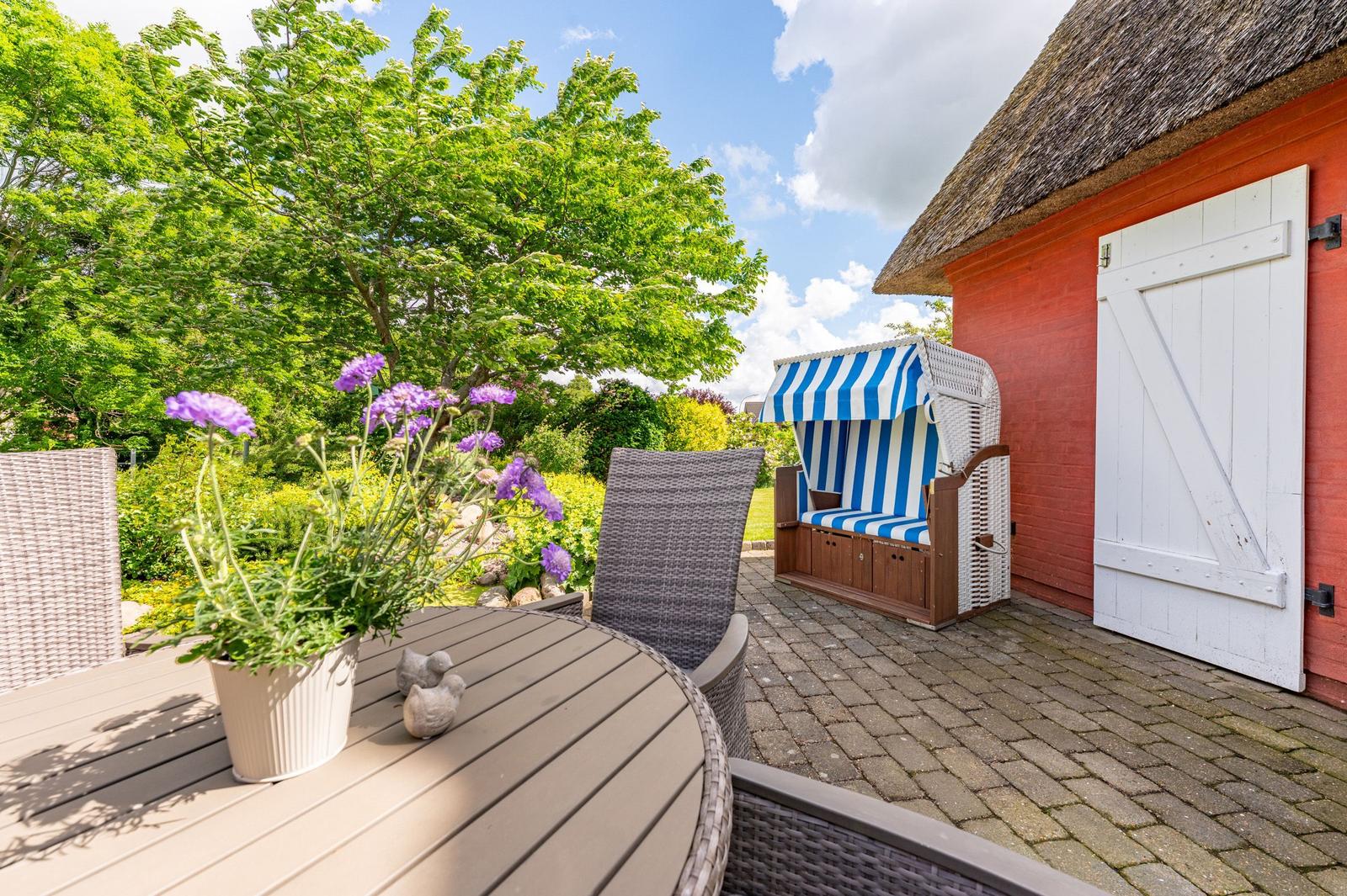 Terrasse mit Tisch, Stühlen und Strandkorb neben rotem Gebäude mit Strohdach.