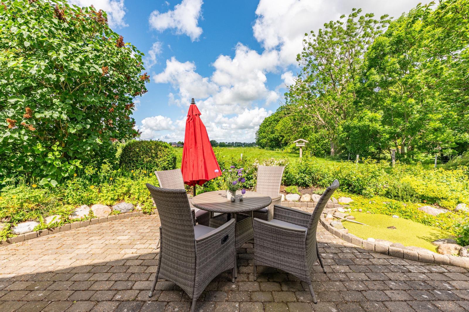 Terrasse mit Tisch, Stühlen und rotem Sonnenschirm. Grünflächen und Bäume im Hintergrund.