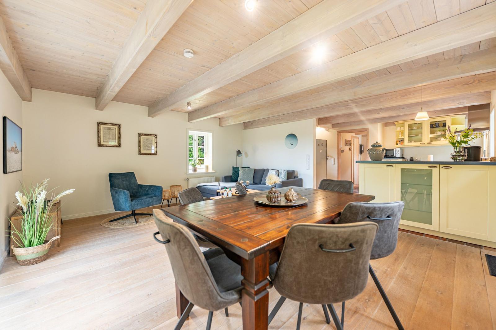 Ein Esszimmer mit Holztisch und Stühlen. Im Hintergrund ist eine Küche und ein Wohnbereich mit Sofa und Sessel.