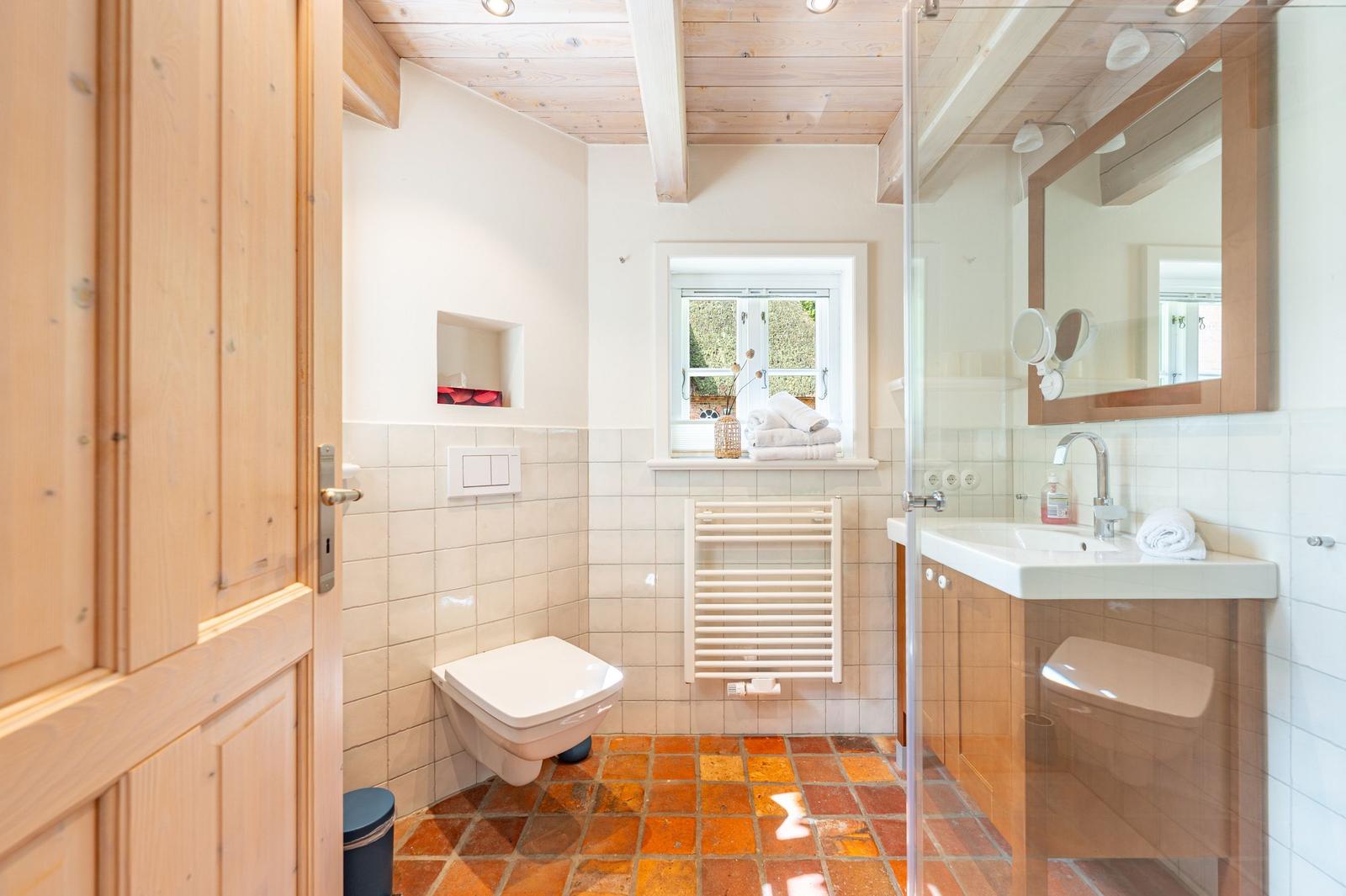 Badezimmer mit Dusche, Toilette und Waschbecken. Holzdecke, Fenster und Terrakottboden.