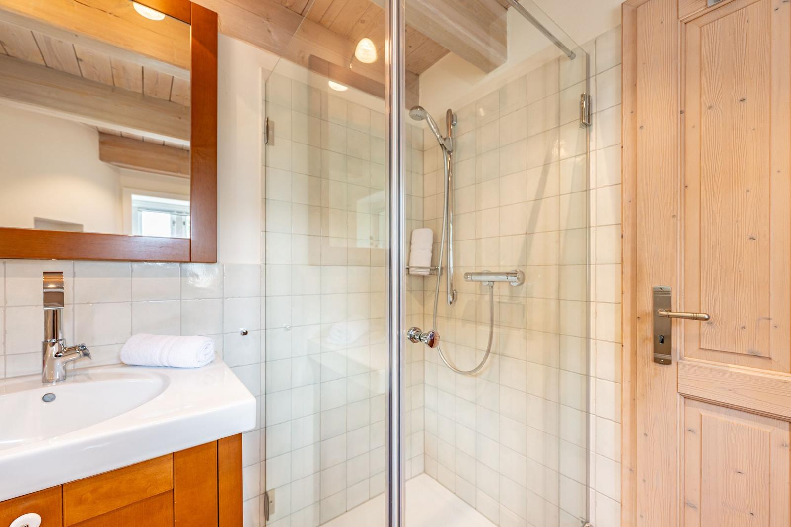 Badezimmer mit Dusche, Waschbecken und Spiegel. Holzdecke und Tür sichtbar.