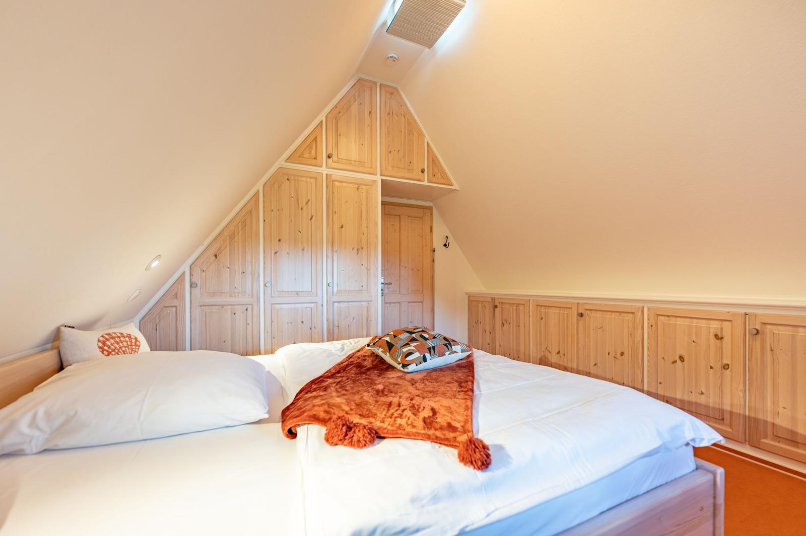 Schlafzimmer mit Dachschrägen, Holzschrank und Bett mit weißer Bettwäsche und orangefarbener Decke.