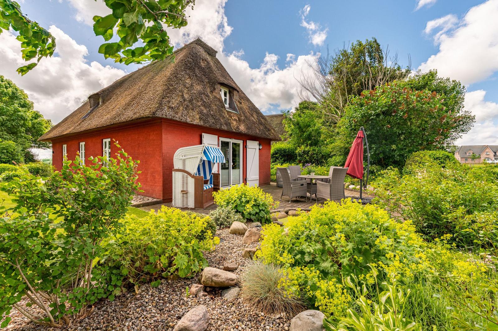 Rotes Haus mit Strohdach, Garten mit Sitzgelegenheiten und Blumenbeeten.