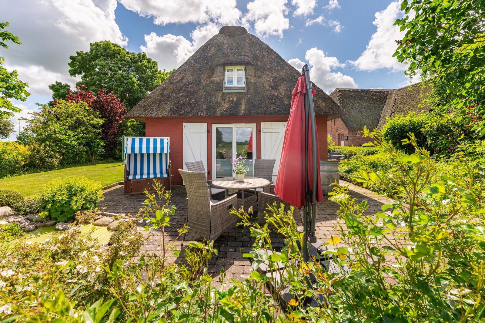Rotes Haus mit Strohdach, Terrasse mit Tisch, Stühlen und Sonnenschirm.