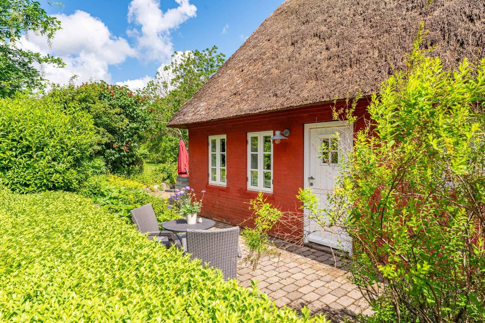 Rotes Haus mit Strohdach, weißer Tür und Fenster. Gartenbereich mit Tisch und Stühlen.