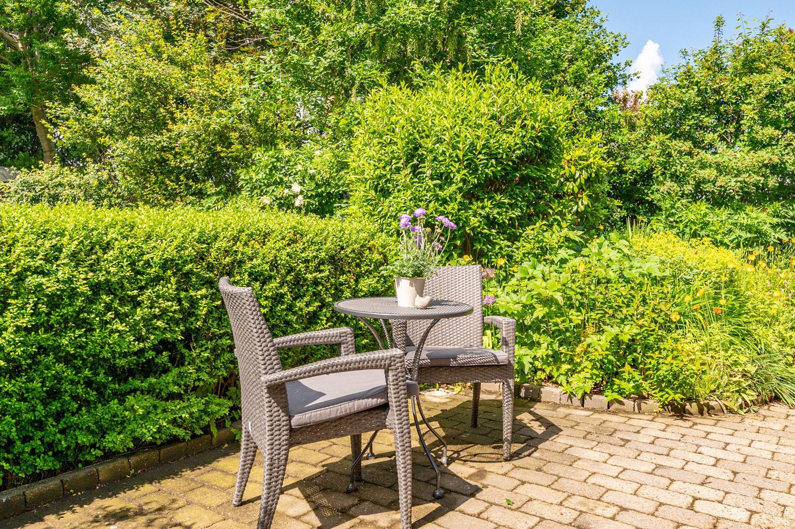 Gartenbereich mit Tisch und zwei Stühlen, umgeben von grünen Sträuchern und Blumen.
