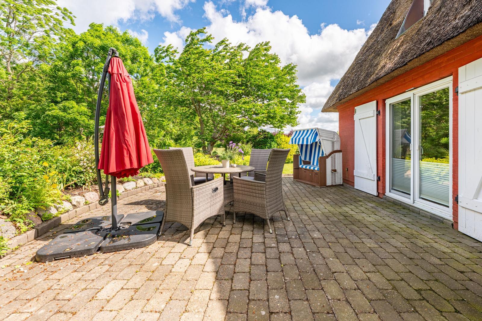 Terrasse mit Tisch, Stühlen und rotem Sonnenschirm neben einem roten Haus mit Dach aus Stroh.