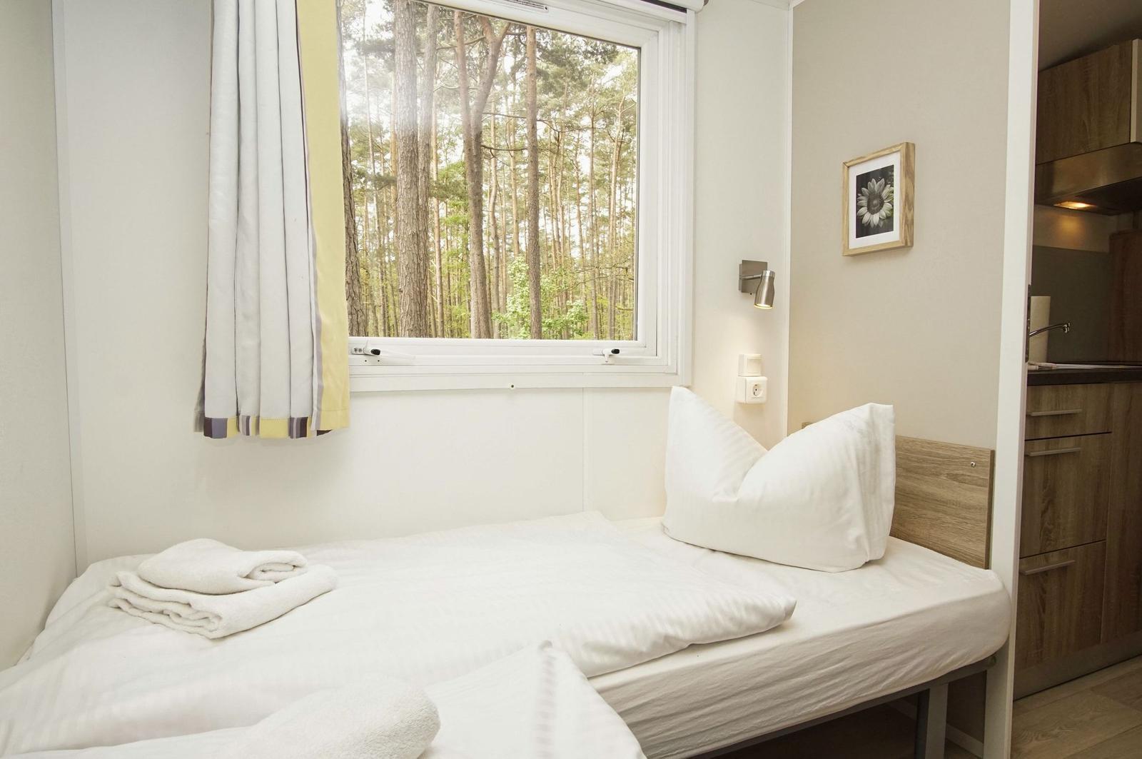 Ein Bett mit weißen Laken und Kissen. Draußen ein Wald. Neben dem Bett ist eine Küchenzeile.