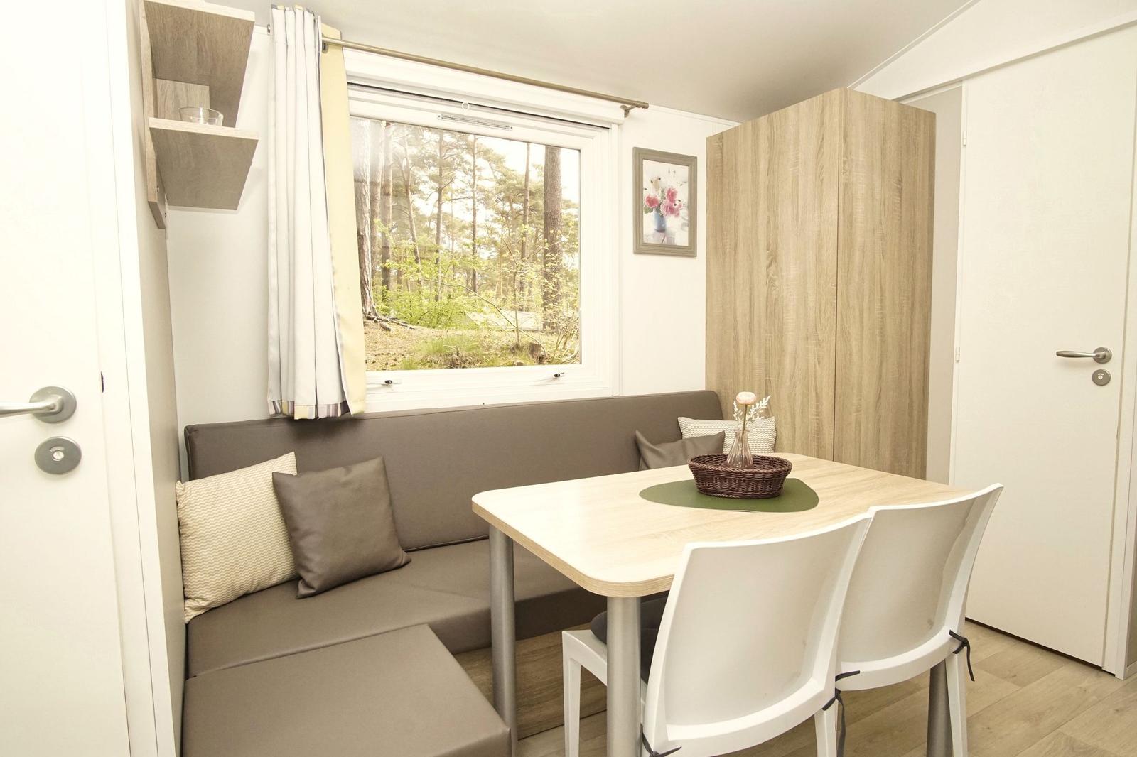 Essbereich mit Sitzbank, Tisch und Stühlen. Fenster zeigt Wald. Schrank und Wandregal sichtbar.
