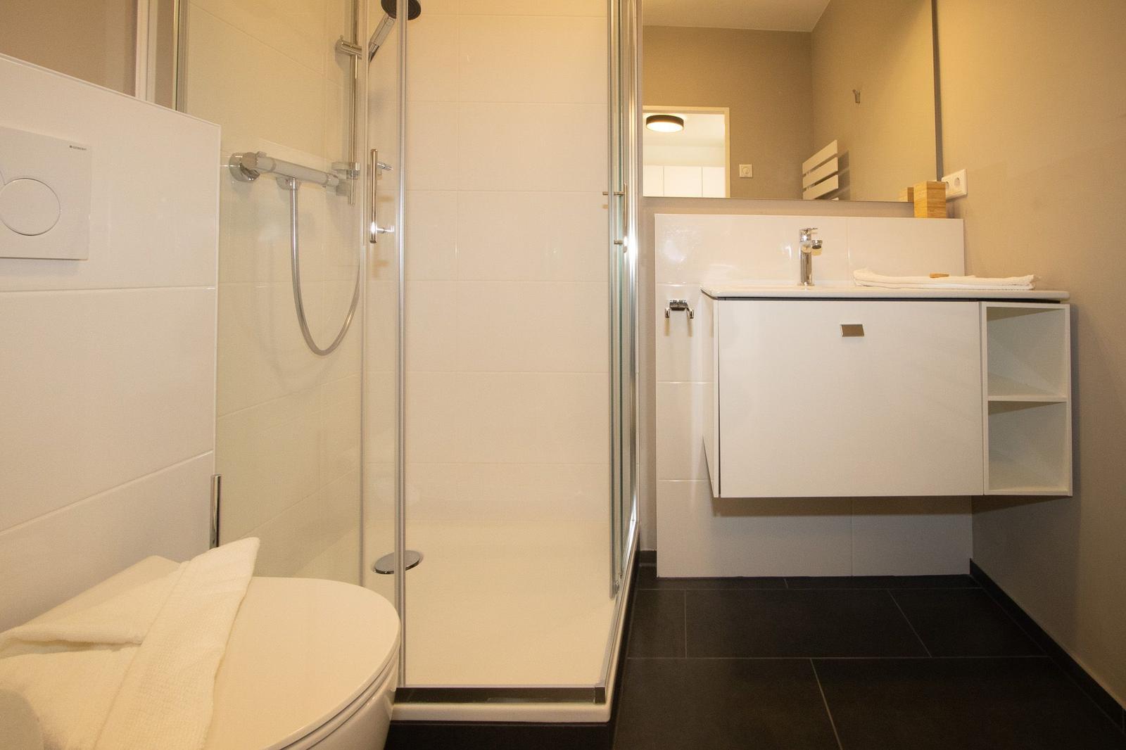 Badezimmer mit Dusche, Waschbecken und Toilette. Weiße Fliesen und dunkler Boden.