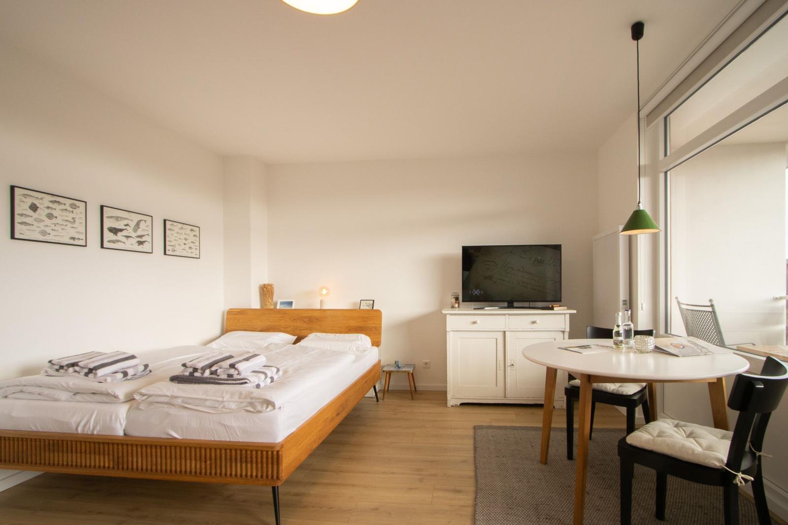 Schlafzimmer mit Bett, Fernseher und Esstisch. Holzboden und große Fenster.