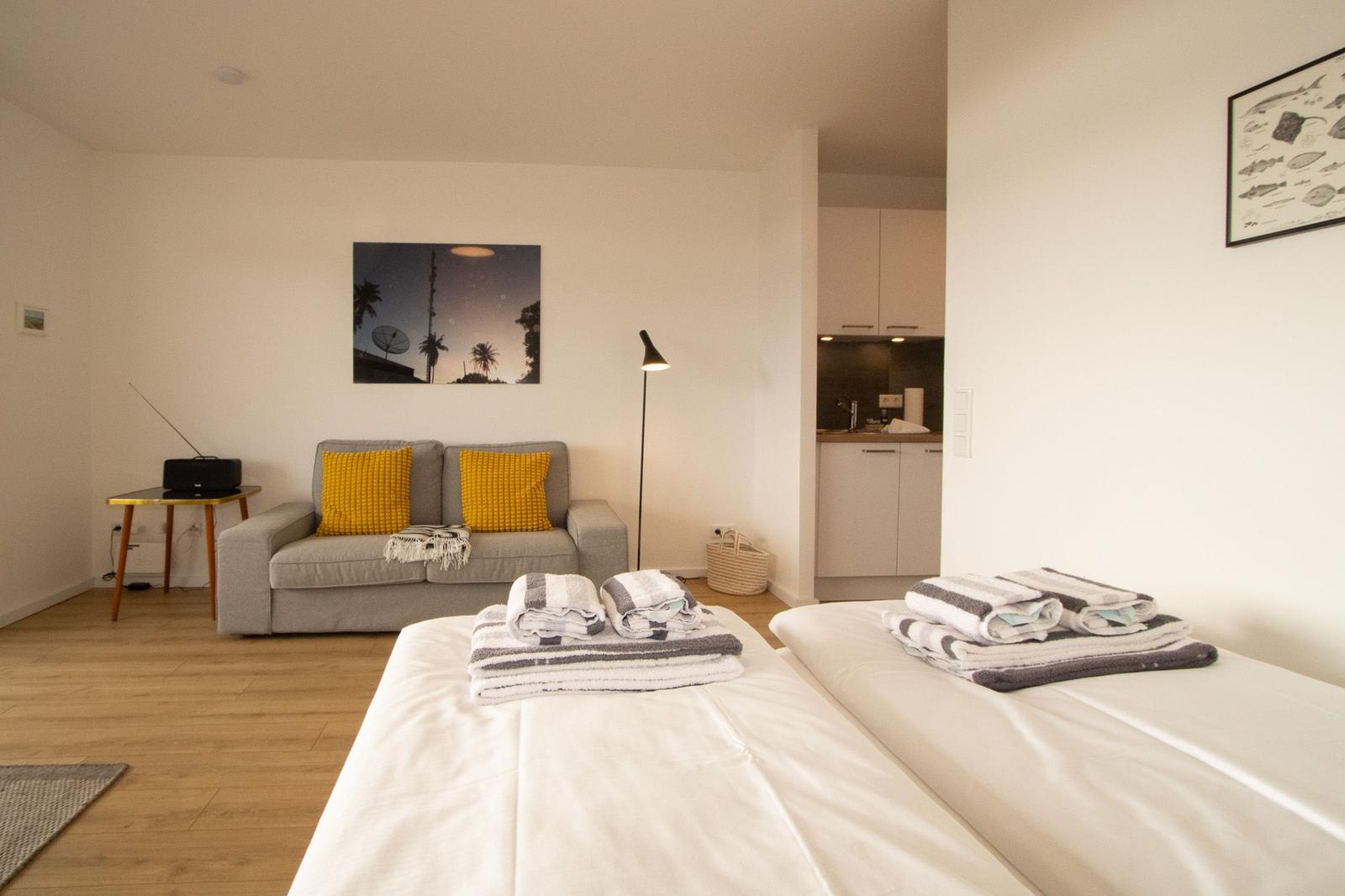 Zwei Betten mit Handtüchern, Sofa mit Kissen, Wandbild und Küchenbereich.