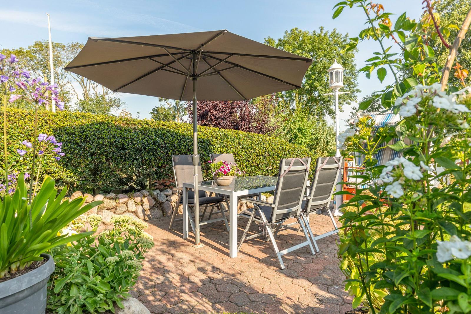 Terrace with table, chairs, and umbrella. Surrounded by flowers and hedge.