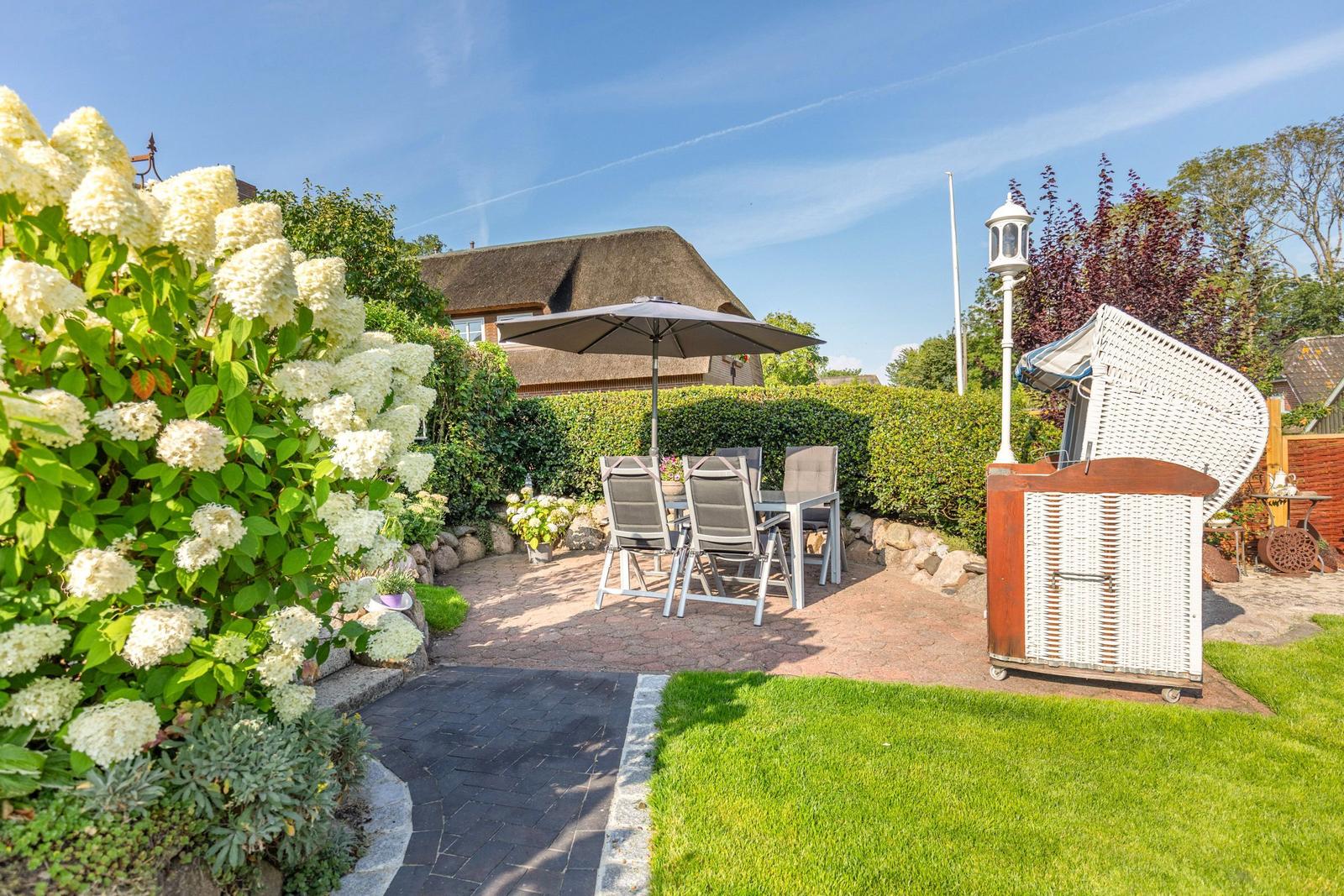 Backyard with seating area, umbrella, and white garden furniture. Lawns and flowers surround the paved area.