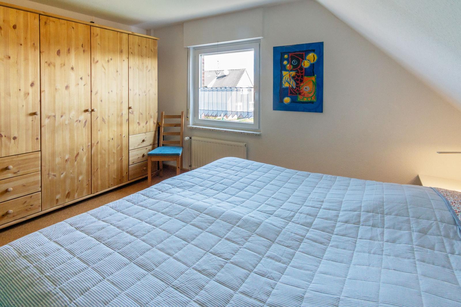 Schlafzimmer mit Doppelbett, Holzschrank und Fenster. Wandbild und Stuhl sind sichtbar.