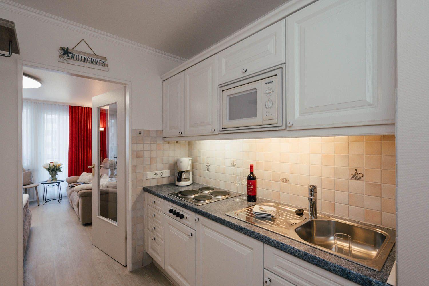 Kitchen with white cabinets, sink, and microwave. Corridor to living area with red curtains.