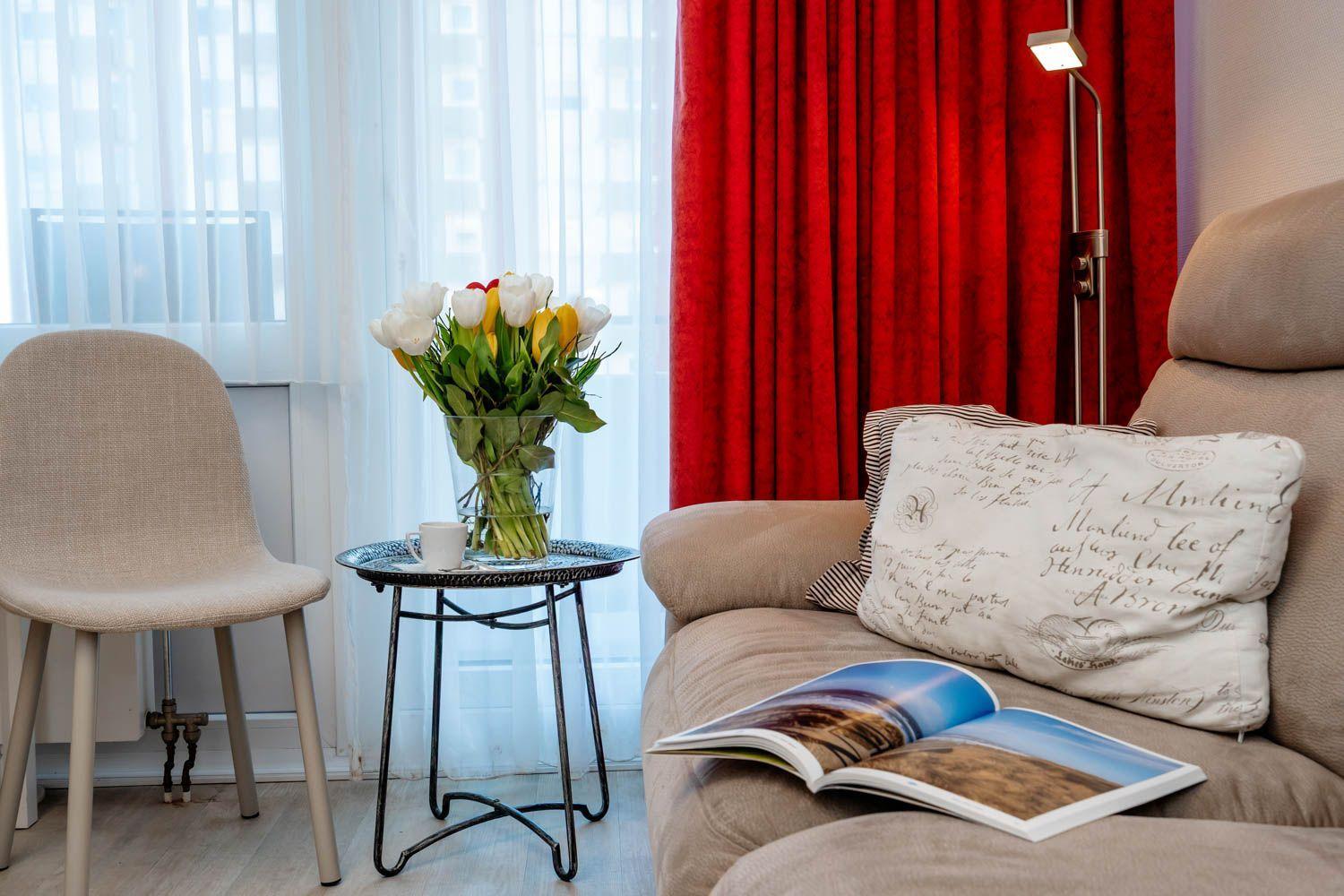 A sofa with cushions and an open book, next to a chair and table with flowers.