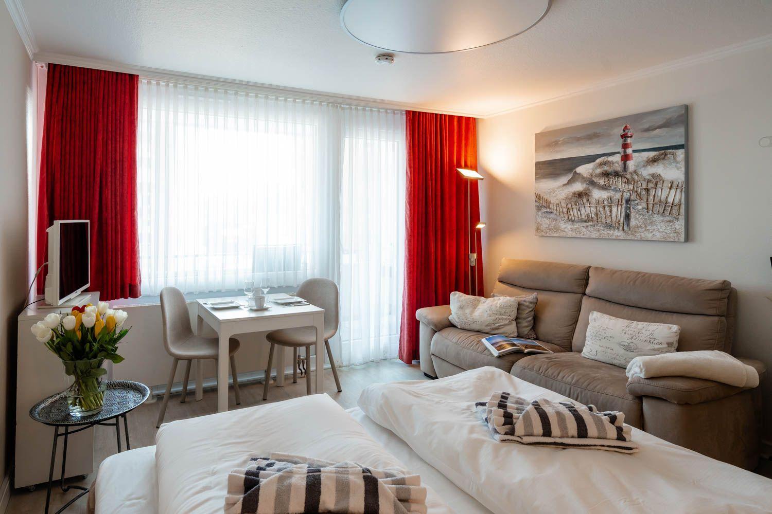 Room with bed, sofa, dining table, and red curtains. Wall art shows lighthouse.