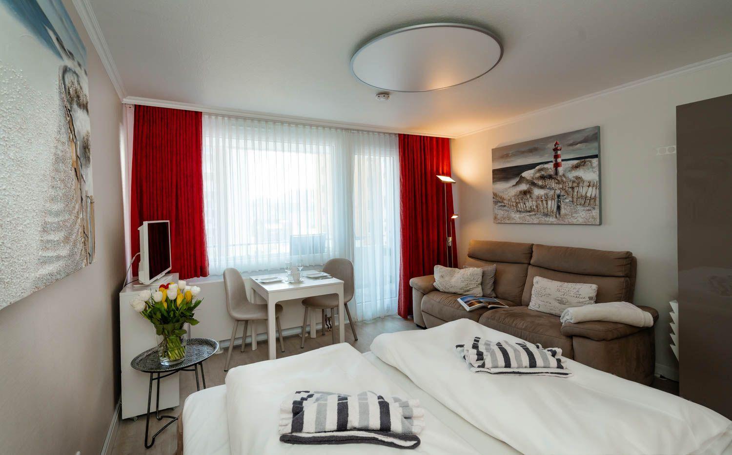 A bedroom with bed, sofa, dining table, and window with red curtains.