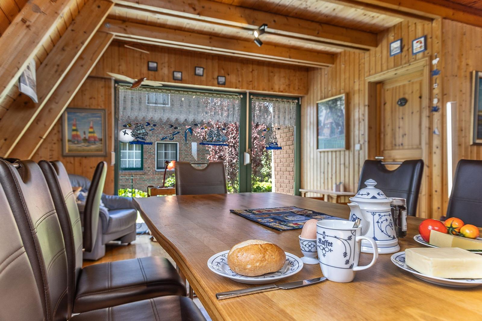 Esszimmer mit Holztisch, Stühlen und Frühstückszubehör. Holzwände und große Fenster.