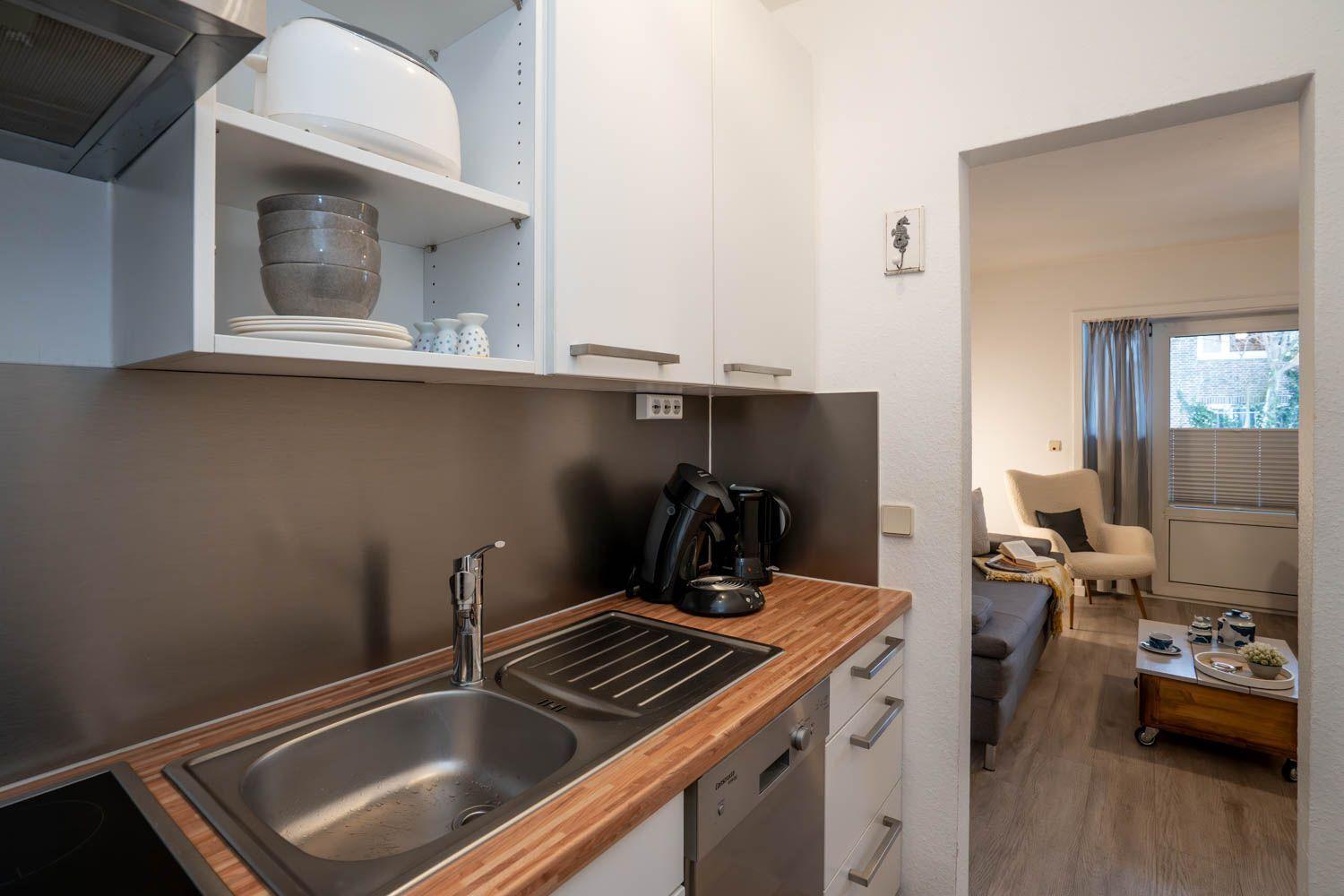 Küche mit Spüle, Arbeitsplatte und Küchengeräten. Durchgang in Wohnzimmer mit Sofa und Fenster.