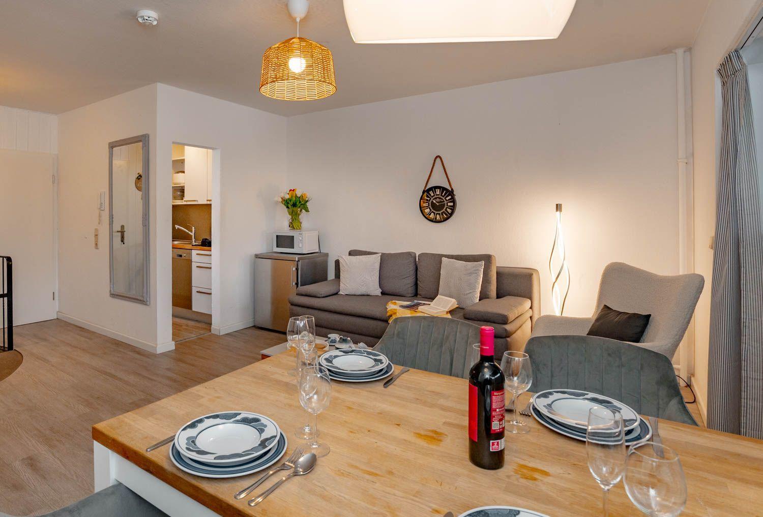 Essbereich mit Tisch, Stühlen und Flasche. Wohnbereich mit Sofa und Küchenbereich im Hintergrund.