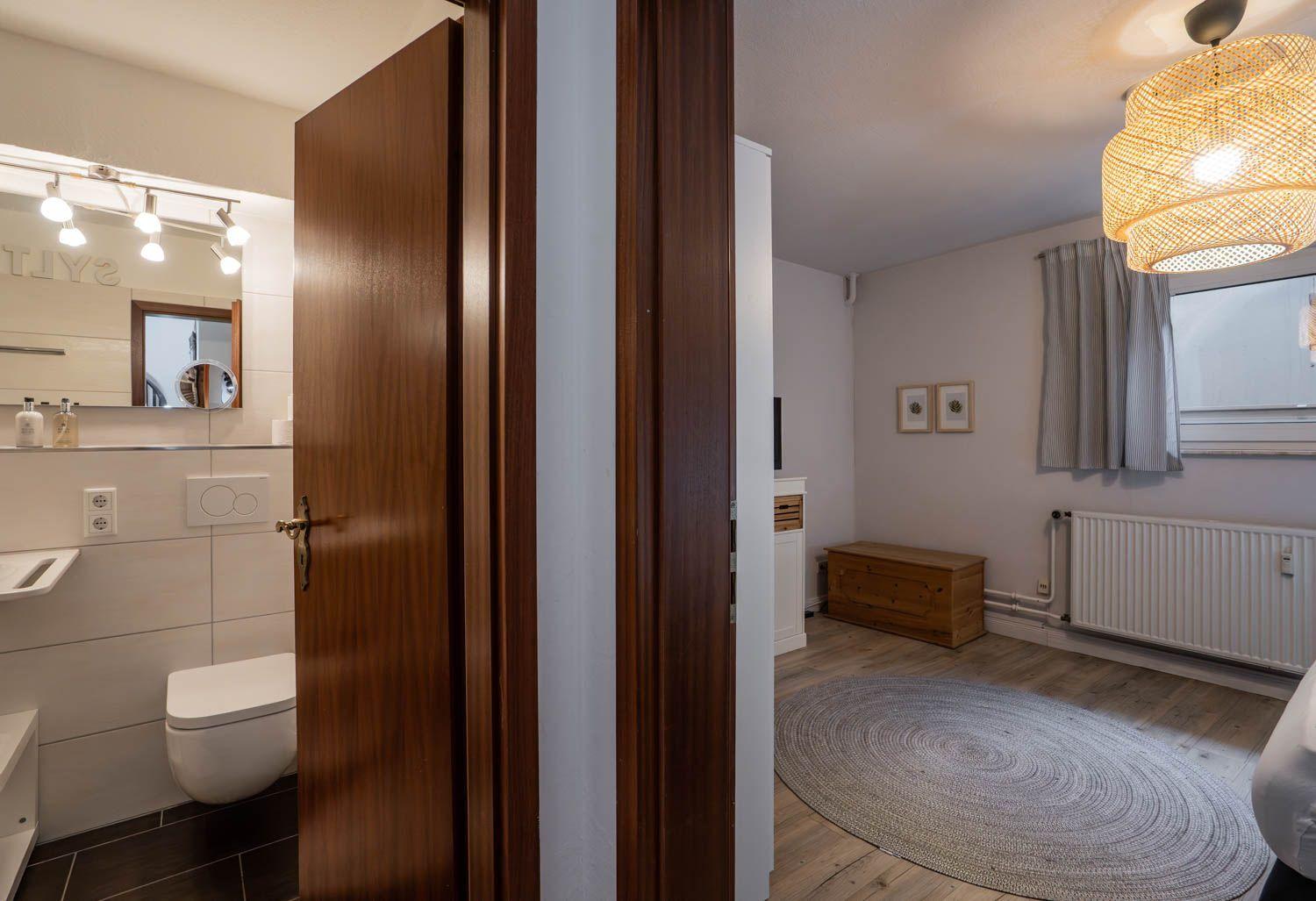 Badezimmer mit Toilette und Spiegelschrank. Nebenraum mit Holzboden, Teppich und Fenster.