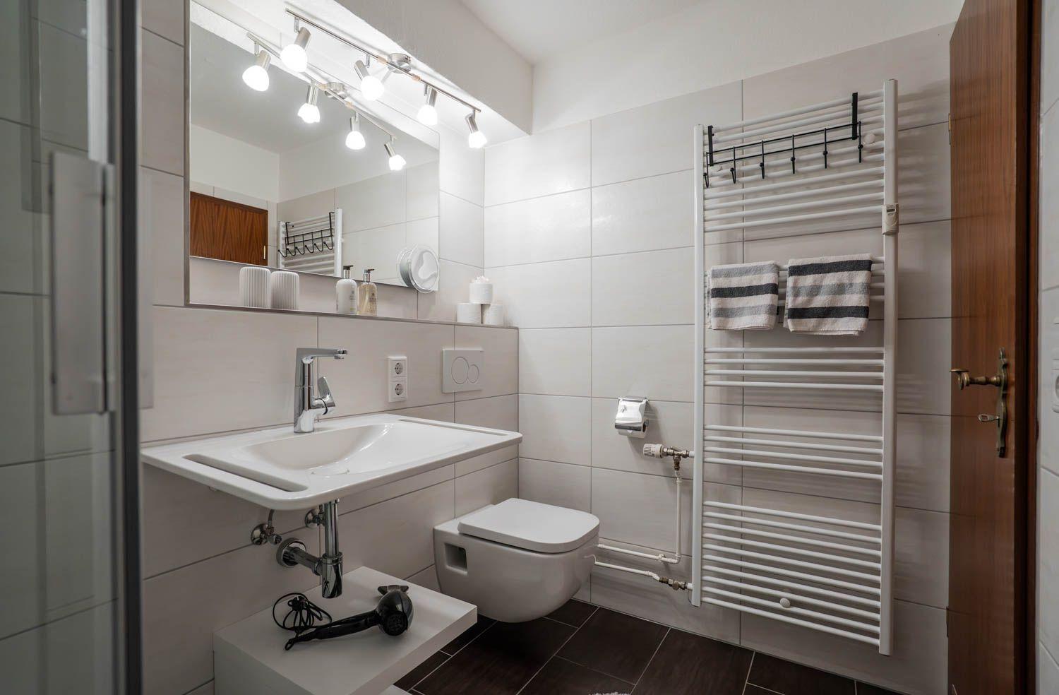 Badezimmer mit Waschbecken, Toilette und Heizkörper. Spiegel mit Beleuchtung und Handtuchhalter.
