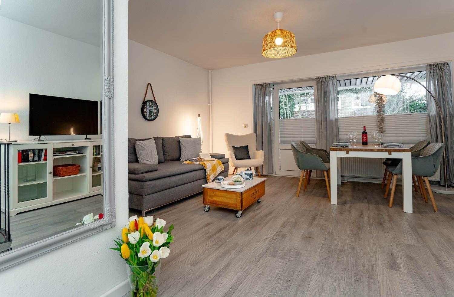 Wohn- und Essbereich mit Sofa, Esstisch und Fernseher. Holzboden und Fenster mit Vorhängen.